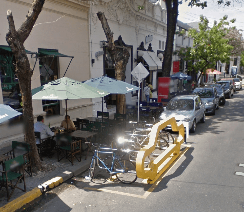 La imagen muestra una vereda con mesas y sillas de un café al aire libre, donde dos personas están sentadas bajo sombrillas verdes de Heineken. Al borde de la acera hay un soporte para bicicletas en forma de contorno de automóvil pintado en color amarillo. Detrás del soporte de bicicletas, se pueden ver varios autos estacionados a lo largo de la calle arbolada, y al fondo, edificios comerciales. La escena refleja un espacio urbano que promueve el ciclismo y el uso del espacio público para peatones y ciclistas en lugar de automóviles.