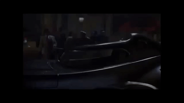 El GIF muestra el batimovil, que parece un automóvil clásico de aspecto elegante y estilizado, en una calle nocturna. La escena está ambientada en un entorno oscuro y urbano, con personas en el fondo que miran el vehículo. La estética y el diseño del auto sugieren un estilo de época o un ambiente noir, evocando misterio o dramatismo.