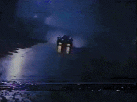 El GIF muestra un automóvil que avanza rápidamente por un camino oscuro y lluvioso. La escena tiene un ambiente sombrío y misterioso, con reflejos de luz en el suelo mojado. La velocidad del auto y la atmósfera nocturna crean una sensación de urgencia y peligro. La iluminación tenue y los reflejos en el agua contribuyen a una estética retro y cinematográfica, posiblemente evocando una escena de una película de ciencia ficción o thriller de los años 80.