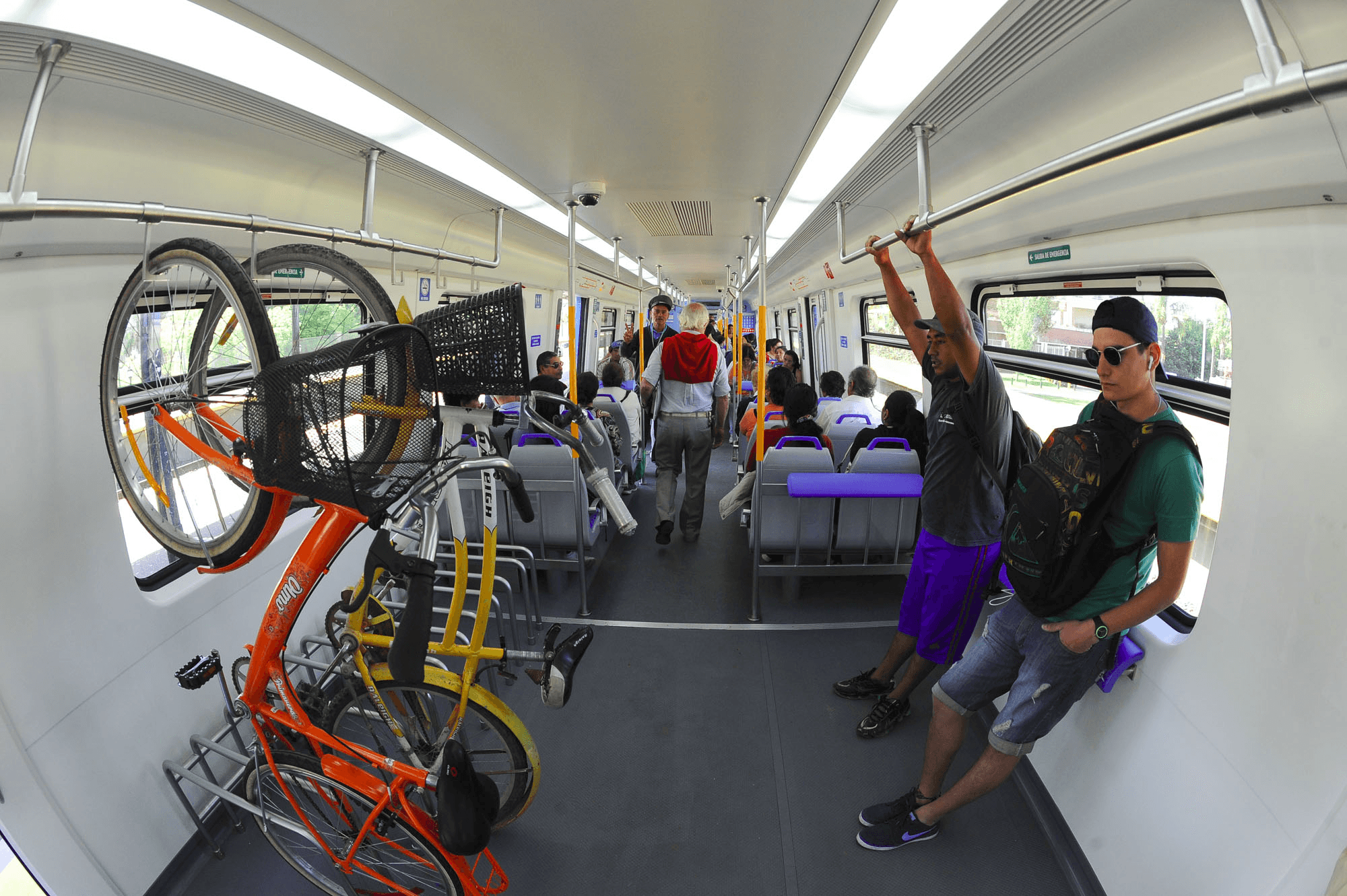La imagen muestra el interior de un vagón de tren con personas sentadas y de pie. En el espacio designado para bicicletas, se observa una bicicleta naranja y otra amarilla colocadas en soportes verticales, facilitando su transporte dentro del tren. Los pasajeros se ven cómodos, algunos de pie sujetándose de las barras de apoyo. La disposición refleja una infraestructura de transporte público amigable con la movilidad en bicicleta, permitiendo a los usuarios combinar ambos medios para sus desplazamientos.