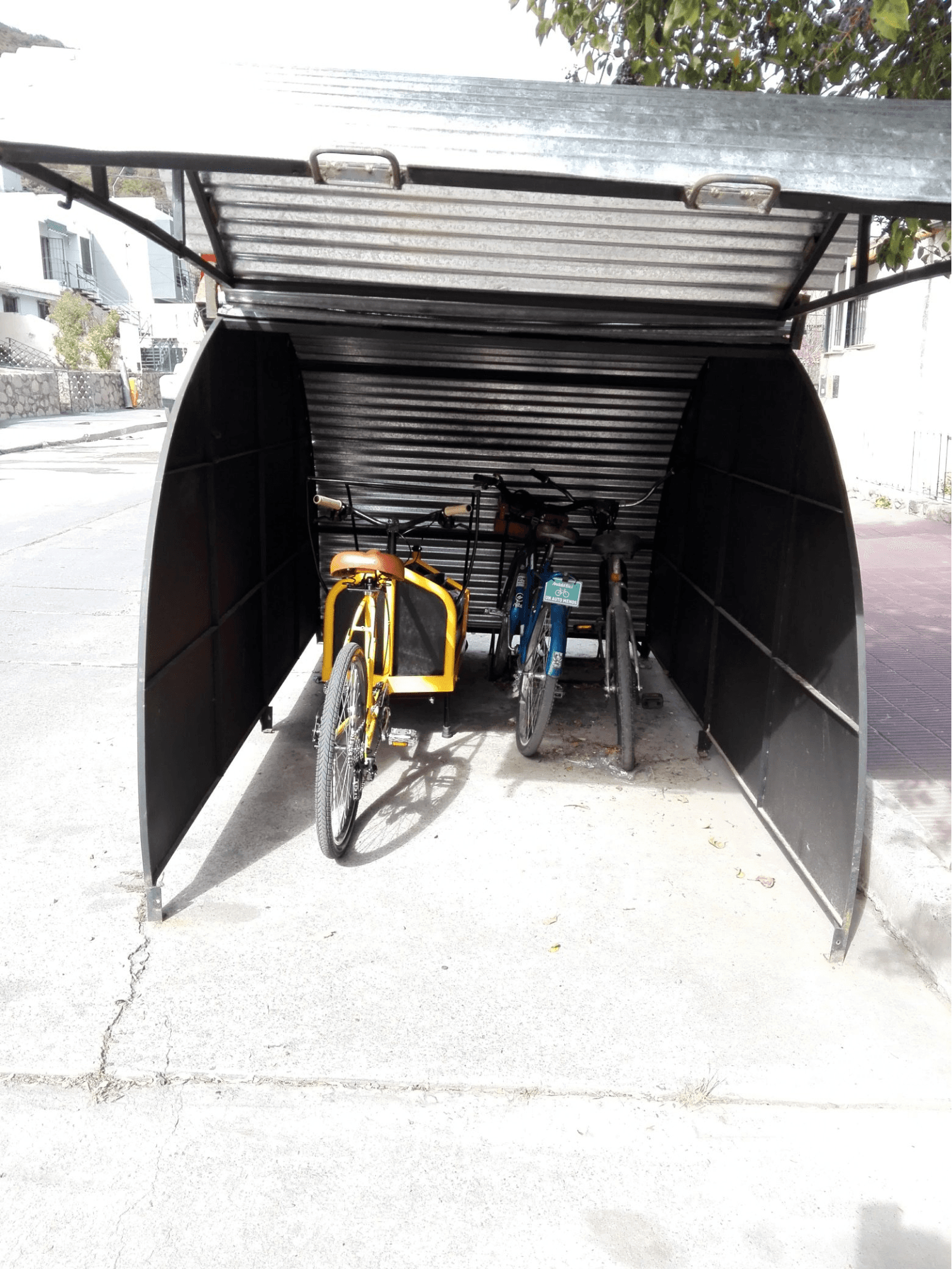 La imagen muestra un refugio cubierto para bicicletas en una vereda. El refugio tiene una estructura metálica semicerrada y un techo corredizo que protege las bicicletas del clima. Dentro del refugio, se pueden ver varias bicicletas estacionadas, incluyendo una bicicleta de carga amarilla. La estructura permite que las bicicletas estén seguras y protegidas, promoviendo el uso de bicicletas al brindar una opción de estacionamiento resguardada.