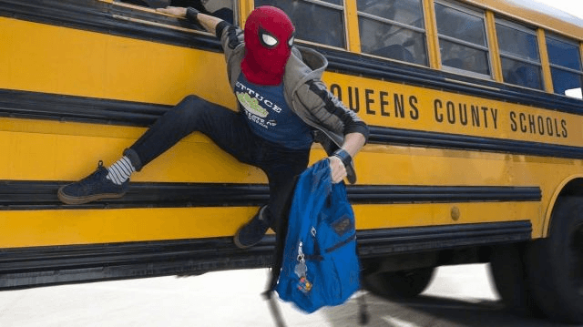 Persona disfrazada de Spider-Man, colgada de un autobús escolar amarillo con el texto 'Queens County Schools'